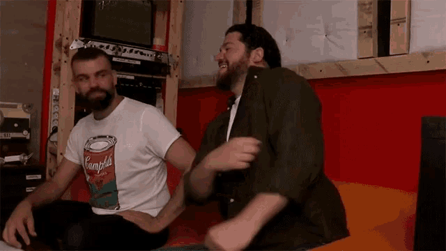 two men are sitting on a couch with one wearing a shirt that says campbell 's soup