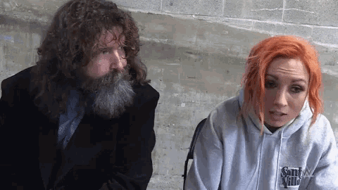 a man with a beard and a woman with red hair are sitting next to each other in front of a wall .