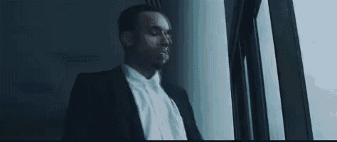 a man in a suit and white shirt is leaning against a wall and looking out a window .