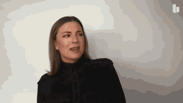 a woman in a black jacket is standing in front of a white wall and making a funny face .