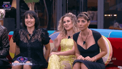 three women are sitting on a couch in front of a sign that says grande fratello on it