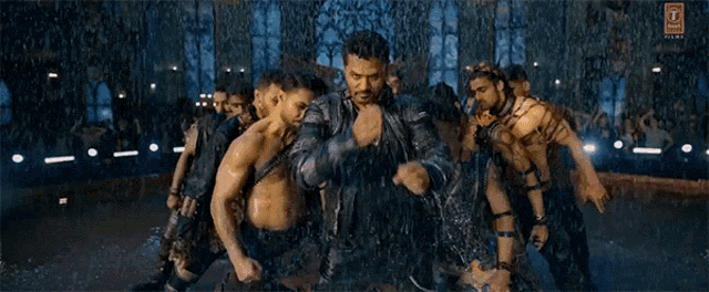 a group of men are dancing in front of a crowd with the letter t on the bottom left