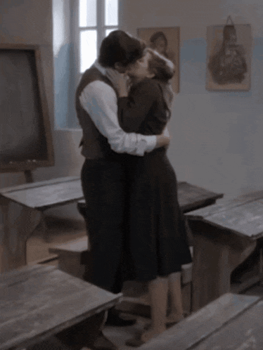 a man and a woman are kissing in a classroom with a blackboard in the background