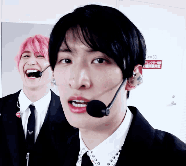 a man wearing a microphone laughs next to another man wearing pink hair