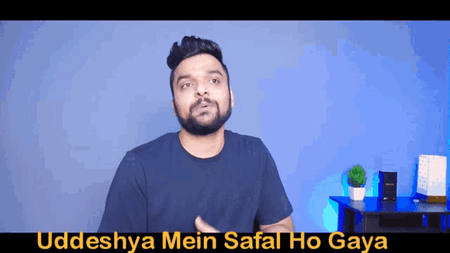 a man with a beard stands in front of a blue wall with the words uddeshya mein safal ho gaya on the bottom