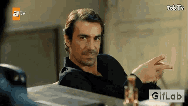 a man in a black shirt is sitting at a table with a bottle of alcohol in front of him .
