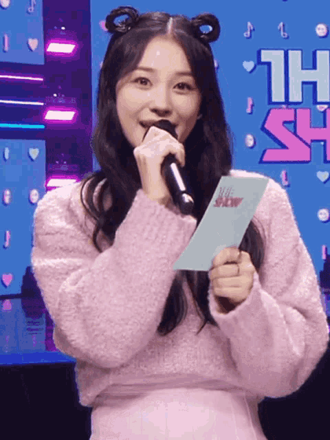 a woman in a pink sweater is holding a microphone and a piece of paper that says show