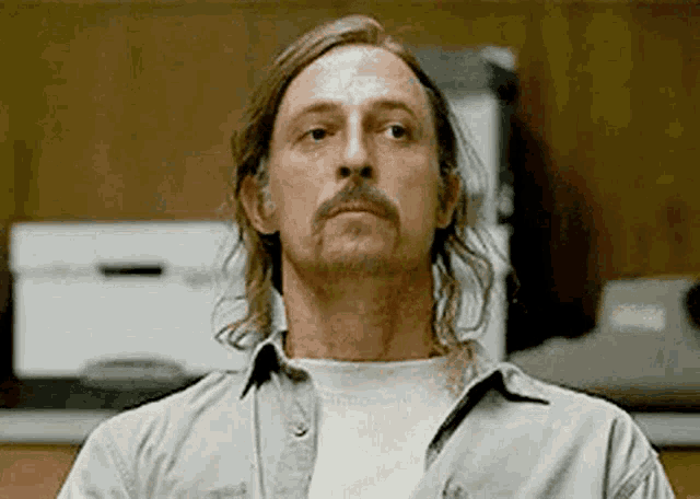 a man with long hair and a mustache is wearing a white shirt and looking at the camera .
