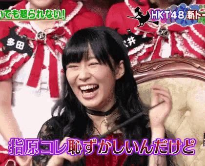a woman is laughing while sitting in a chair in front of a group of hkt48 members .