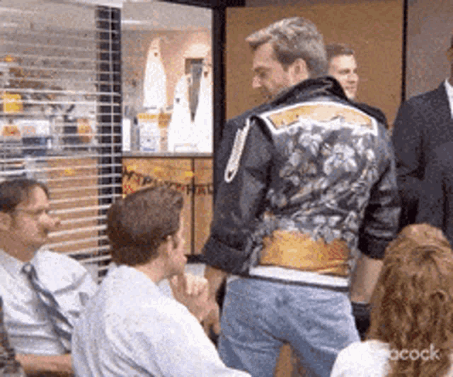 a man in a motorcycle jacket is standing in front of a group of people in an office .