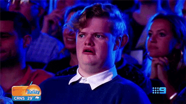 a man is making a surprised face while sitting in a crowd of people watching a tv show .