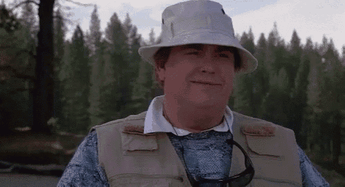 a man wearing a hat , vest and sunglasses is standing in front of a forest .