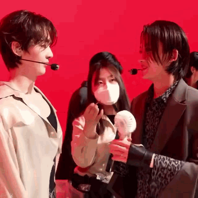 two men are standing next to each other in front of a red background while a woman wearing a mask holds a fan .