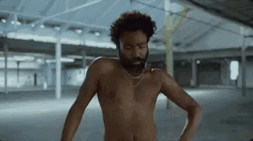 a shirtless man with a beard and a necklace is standing in a warehouse .