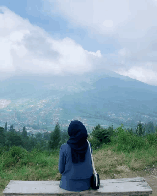 a woman in a blue hijab sits on a bench overlooking a mountain
