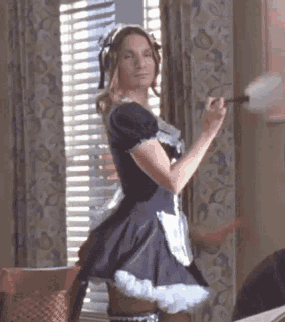 a woman dressed as a maid is standing in front of a window holding a duster .