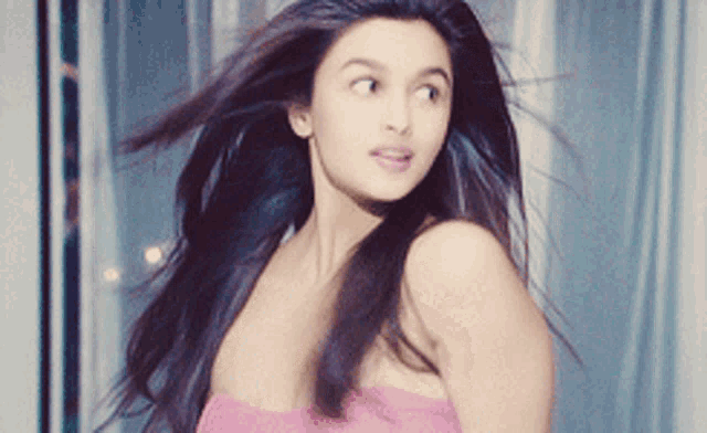 a woman in a pink strapless top is standing in front of a window with her hair blowing in the wind