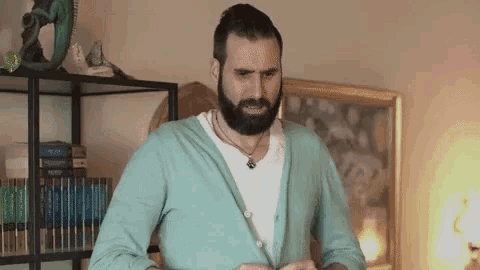 a man with a beard wearing a blue cardigan is standing in front of a bookshelf .