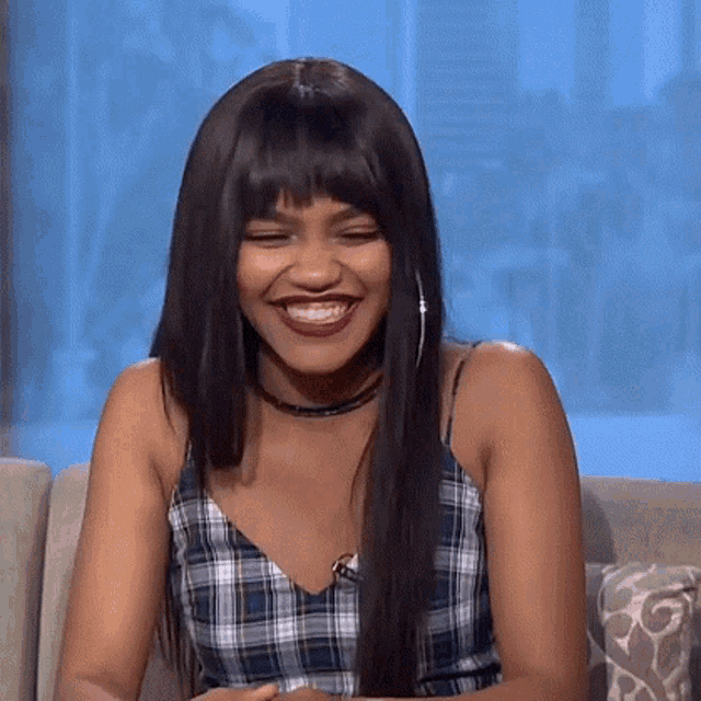 a woman is smiling while sitting on a couch wearing a plaid top and choker .