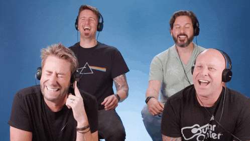 four men wearing headphones are laughing and one has a shirt that says guitar killer