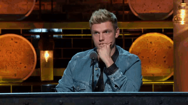 a man in a denim jacket is sitting in front of a microphone with his hand on his chin