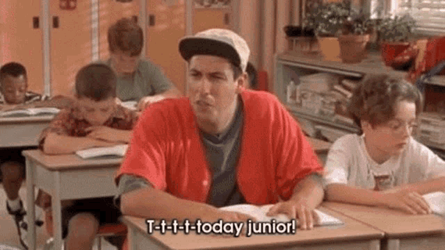 a man in a red shirt is sitting at a desk in a classroom talking to a boy .