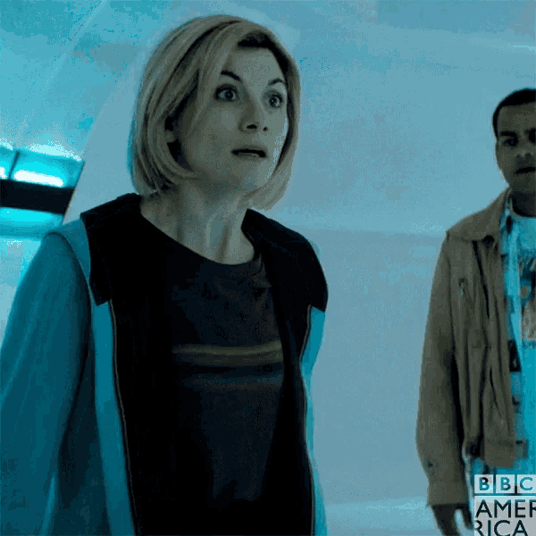 a woman in a blue jacket is standing next to a man in a brown jacket with bbc america written on the wall behind her
