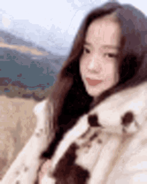 a woman in a white coat is taking a selfie in a field with mountains in the background .