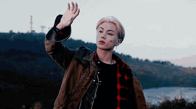 a young man wearing a plaid shirt and a brown jacket is standing in front of a body of water
