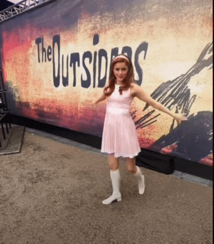 a woman in a pink dress is standing in front of a sign for the outsiders