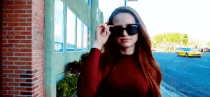 a woman wearing sunglasses is standing next to a brick building .