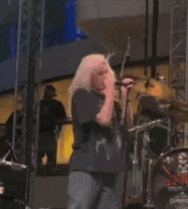 a woman is singing into a microphone while standing on a stage in front of a drum set .