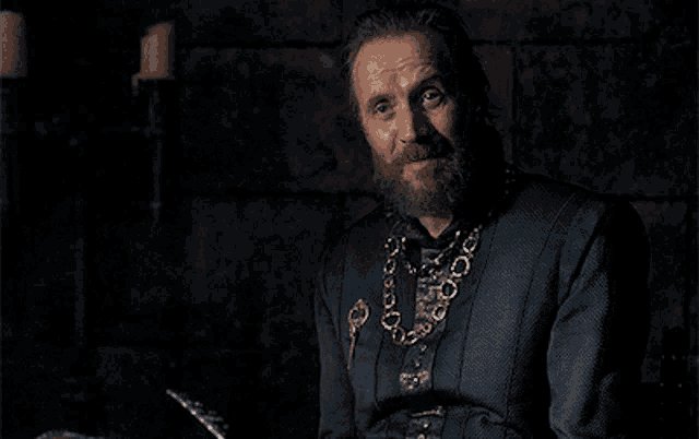 a man with a beard and a chain around his neck is sitting in a dark room .