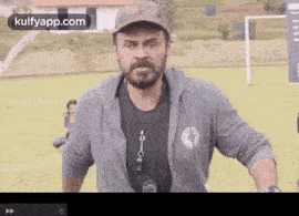 a man with a beard is standing on a soccer field wearing a hat and a hoodie .