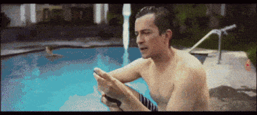 a shirtless man sits by a pool looking at his cell phone