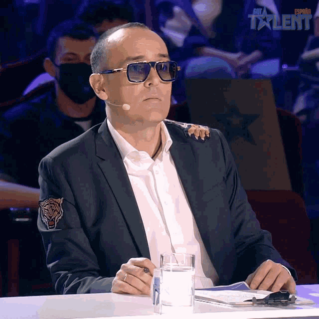 a man in a suit sits at a table with a glass of water in front of a sign that says espana talent