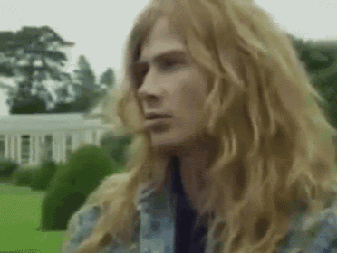 a man with long blonde hair and a blue shirt is standing in a field .