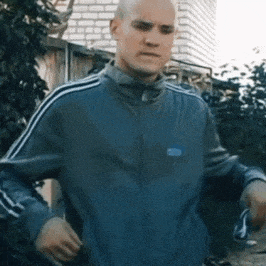 a bald man wearing a grey adidas jacket stands in front of a brick building