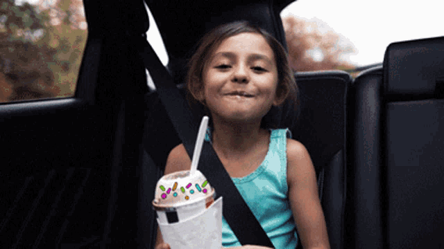 a little girl sitting in a car seat holding a cup of milkshake with sprinkles on it