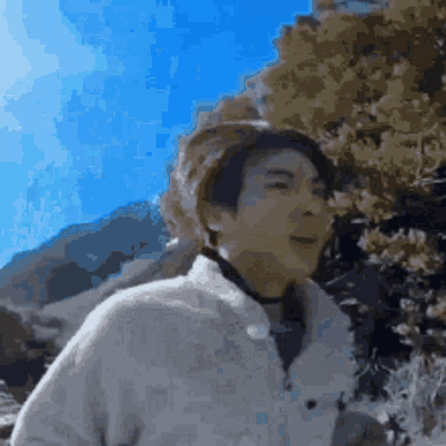 a man in a white sweater is standing in front of a mountain with trees in the background .