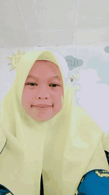a girl wearing a yellow hijab smiles in front of a sign that says 12uh