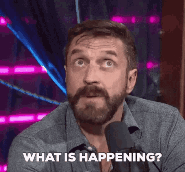 a man with a beard and mustache is sitting in front of a microphone and asking what is happening .