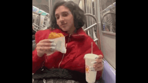 a woman is sitting on a subway train eating a sandwich and drinking a cup of soda .