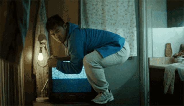 a man in a blue jacket is squatting down in front of a flat screen tv