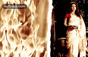 a woman in a white saree is standing in front of a wall of fire .