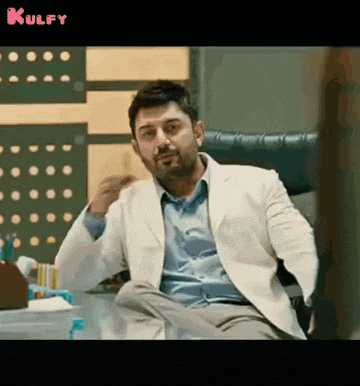 a man in a white jacket is sitting at a desk and talking to someone .