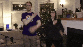 a man and woman are clapping in front of a christmas tree in a living room