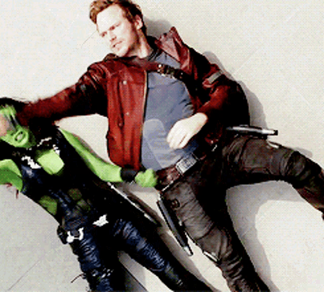 a man in a red leather jacket is laying next to a woman in a green costume