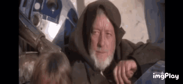 a man with a beard and a hooded jacket is sitting next to a robot .