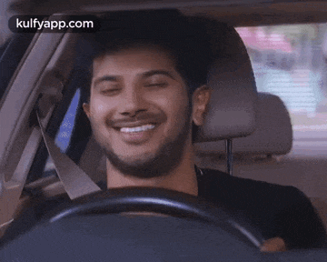 a man is smiling while driving a car and the website kulfyapp.com can be seen in the corner